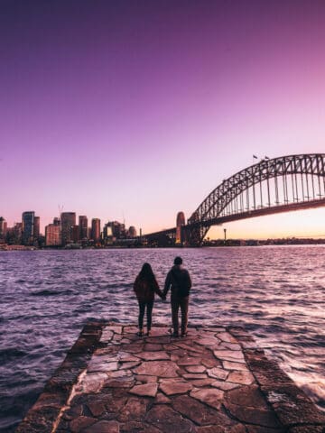 A couple holds hands by the water during a purple sky sunset in Sydney (photo: EyeEm Mobile GmbH, iStock).