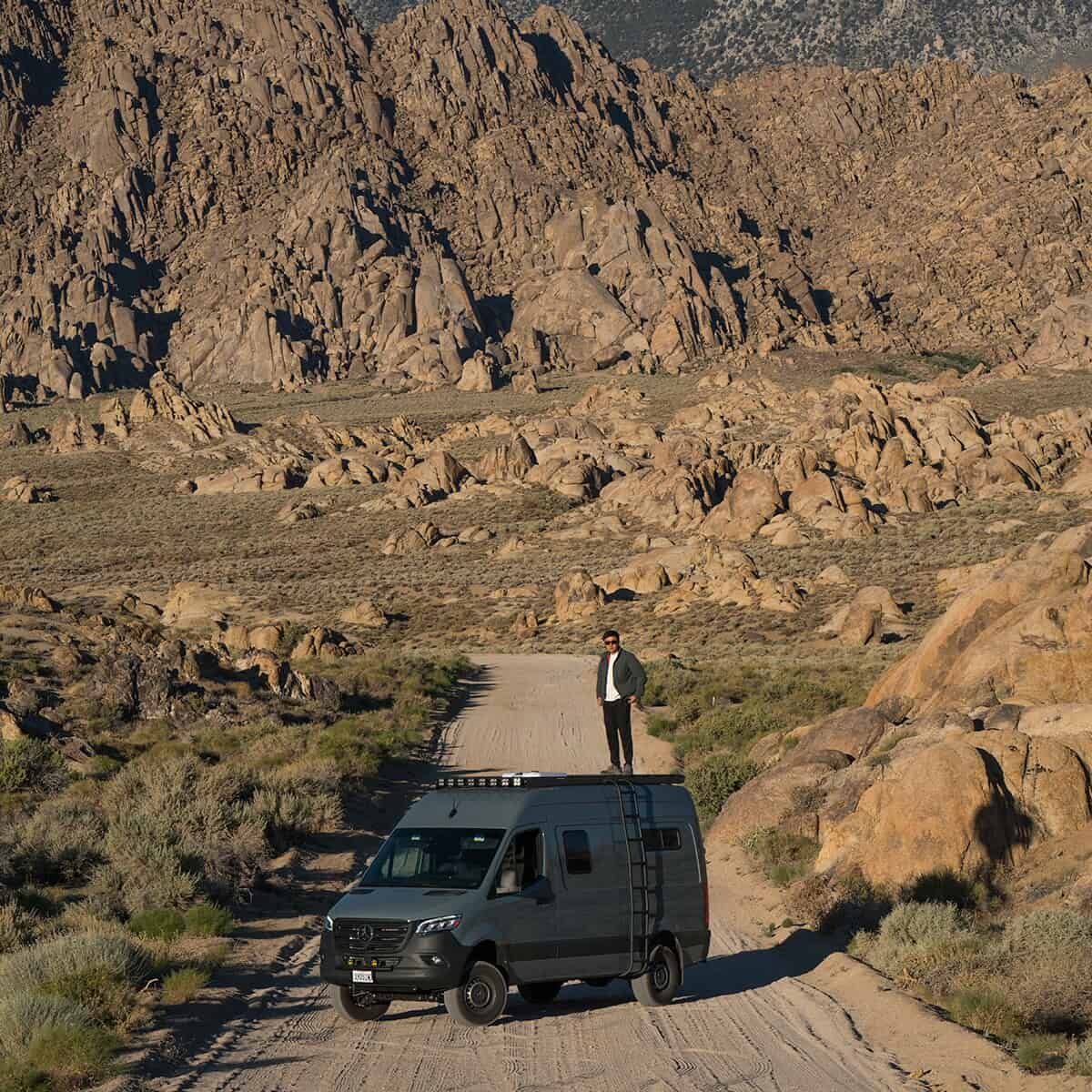 Man standing on a grey Sprinter van on a dirt road with a rocky landscape.