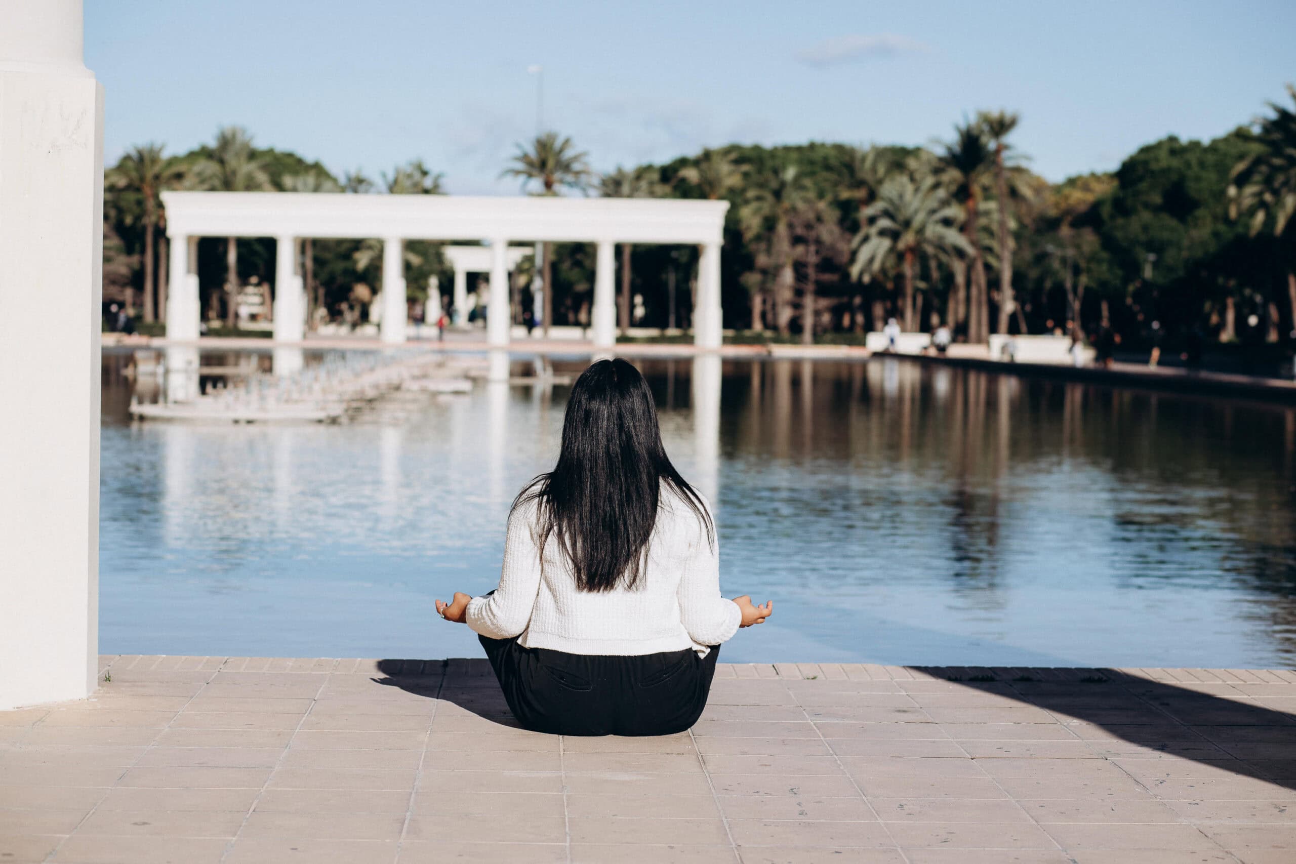 A serene scene in Turia Gardens in Valencia, capturing a woman sitting by the water surrounded by palm trees and modern architectural elements, reflecting the calm and beauty of one of the city's most iconic parks.
