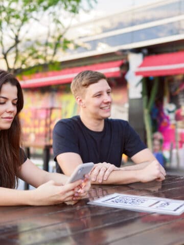 Travelers using a smartphone to access a QR code menu at an outdoor restaurant.