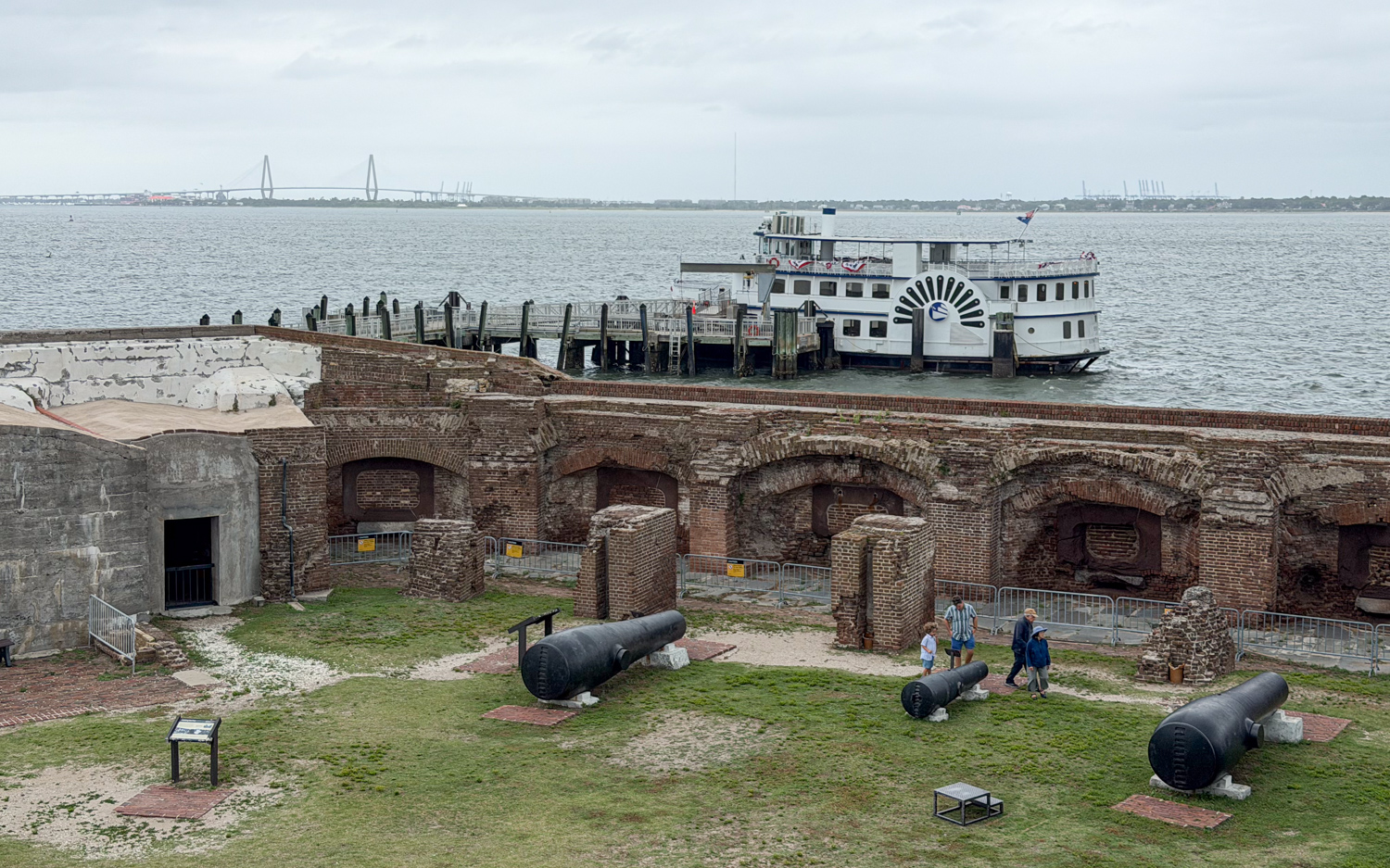 Cannons and brick ruins inside Fort Sumter with a tour boat docked beyond the wall.