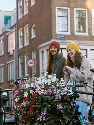 Two women with bicycles on a flower-lined bridge in Amsterdam