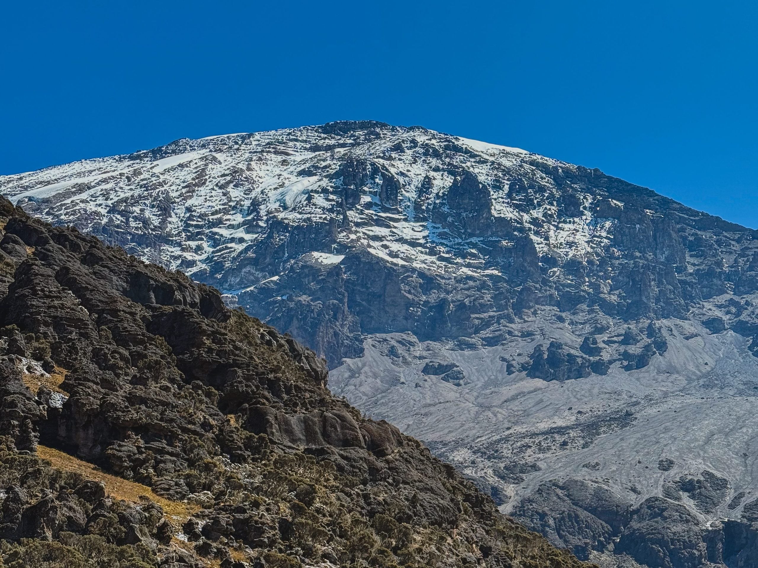 Snow-streaked cliffs on Kilimanjaro above a rocky foreground under a blue sky.