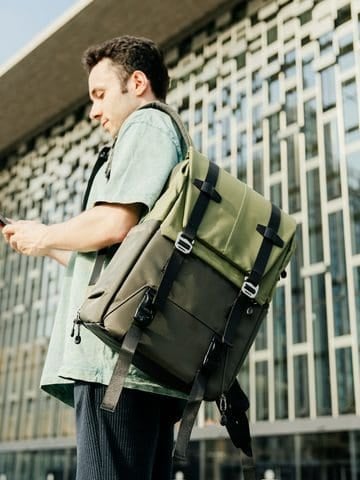 Man wearing a green backpack checks his smartphone outside a modern building.