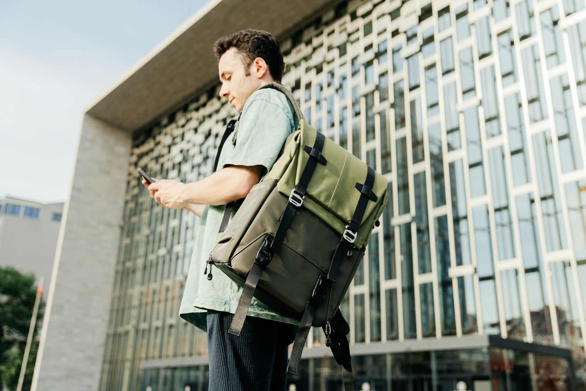 Man wearing a green backpack checks his smartphone outside a modern building.